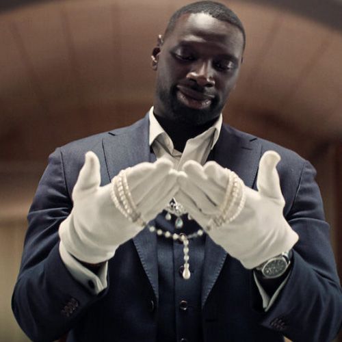 Omar Sy, con una expresión pensativa, sostiene un collar de perlas blanco en sus manos enguantadas de blanco. Lleva un traje azul oscuro y una camisa blanca desabrochada.